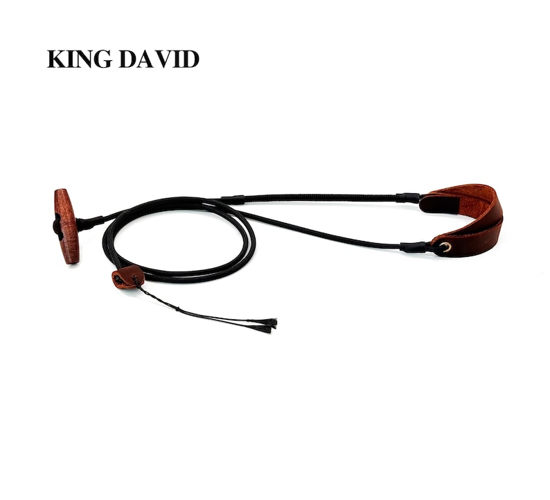 May include: A black cord strap with a brown wooden toggle and brown leather accents. The text KING DAVID is printed in black in the upper left corner. The strap features a loop design with a sliding leather adjustment piece and two thin cord ends.