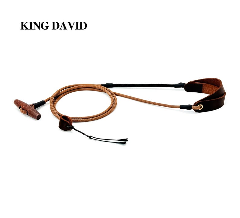 May include: A camera strap labeled KING DAVID featuring a tan cord, a dark brown leather wrist loop, and a wooden toggle fastener. The strap includes black connectors and a thin black tether cord attached to a small leather loop for secure attachment.