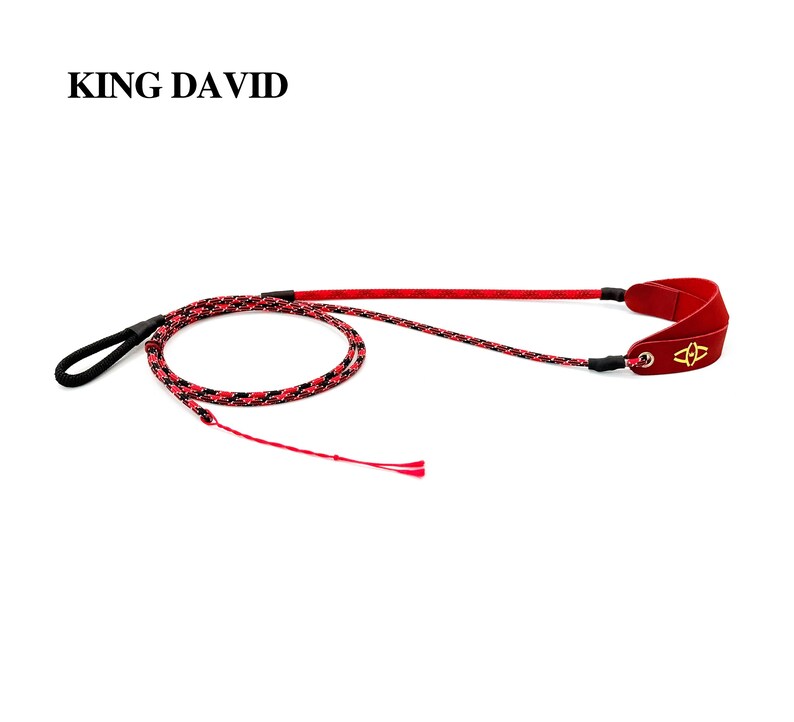 Puede incluir: Una honda de la marca King David con un cord&oacute;n trenzado en rojo y negro, un lazo negro en un extremo y una bolsa de cuero roja en el otro. La bolsa de cuero roja tiene un emblema dorado y est&aacute; sujeta al cord&oacute;n mediante un ojal met&aacute;lico.