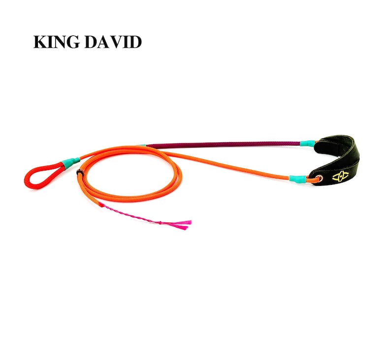Puede incluir: Una honda de la marca King David con un cord&oacute;n naranja brillante, un asa de bucle, una secci&oacute;n morada y una bolsa de cuero negra con un logotipo dorado. El cord&oacute;n est&aacute; asegurado con detalles en azul claro y un extremo de cord&oacute;n rosa deshilachado.