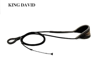 May include: A black sling with the text KING DAVID printed above it. The sling features a long, thin cord, a braided loop at one end, and a wider, padded, curved section at the other end, constructed with visible stitching along the edges.