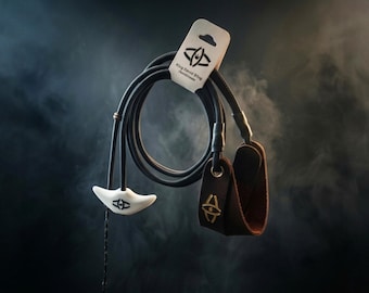 May include: A black cord sling with a white molded release tab and a brown leather finger loop. The product is held by a branded cardboard tag labeled King David Sling. The design features a black logo on the white tab and a gold logo on the leather.
