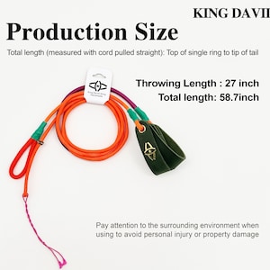 Puede incluir: Una honda hecha a mano King David con cuerda naranja, un lazo de cuero verde oscuro para el dedo con un logo dorado y una cola rosa. El texto indica una longitud de lanzamiento de 68,58 cm y una longitud total de 149,1 cm. Incluye una etiqueta.