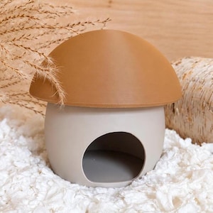 May include: A mushroom-shaped hideout featuring a smooth, rounded tan cap and a light grey base with a large, circular opening in the front. The structure has a clean, matte finish and a hollow interior, designed as a small shelter for small pets.