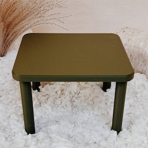 May include: A small, olive green square table with rounded corners and four cylindrical legs. The table is set against a neutral background with decorative elements.