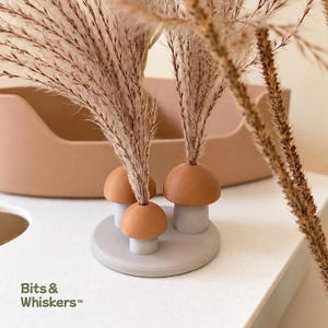 Puede incluir: Un soporte decorativo con tres soportes en forma de seta sobre una base circular, con sombreros de color marrón claro y tallos de color gris claro. Tallos de hierba de la pampa seca están insertados en las setas. Aparece el texto Bits & Whiskers.