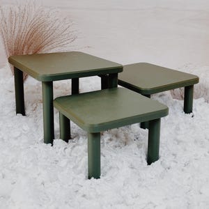 Peut inclure: Un ensemble de trois tables vert olive avec des plateaux carrés et quatre pieds cylindriques chacune. Les tables sont disposées à des hauteurs variables, présentant des surfaces lisses et plates avec des coins arrondis.