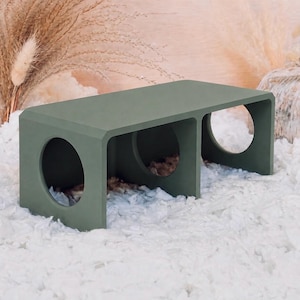 May include: A rectangular olive green pet tunnel featuring three circular openings along the front panel. The structure has a smooth, matte finish and is designed with a hollow interior, providing a clear view through each of the three circular cutouts.