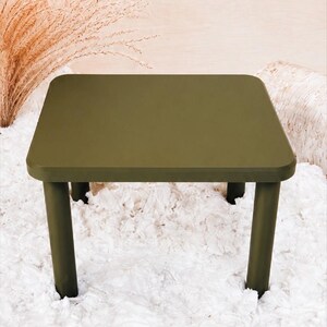 May include: A square, olive green table with rounded corners and four thick, cylindrical legs. The table features a smooth, matte surface finish and a simple, clean design with no additional ornamentation or visible hardware on the exterior.