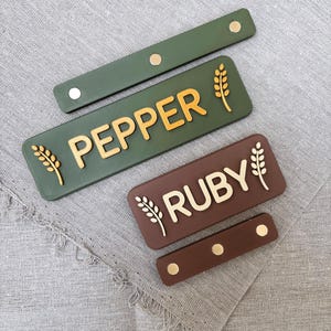 Puede incluir: Dos etiquetas rectangulares en verde oliva y marrón oscuro, con los nombres PEPPER y RUBY en letras doradas en relieve. Cada etiqueta está flanqueada por iconos de espigas de trigo doradas y acompañada de una correa de cuero a juego con tres remaches dorados.