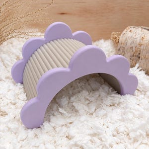 Puede incluir: Un juguete en forma de arco semicircular con dos paneles laterales festoneados de color lavanda y una serie de barras horizontales paralelas de color beige claro que los conectan. La estructura tiene un acabado mate y liso.