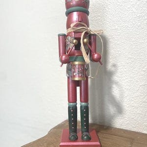 May include: A decorative wooden nutcracker figurine in shades of red, green, and gold. The figurine has a drum, boots, and a hat with floral accents. A beige ribbon is tied around the waist. The nutcracker stands on a red base.