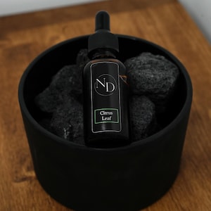 May include: A dark amber glass dropper bottle with a black cap and a black label featuring the letters ND and the text Citrus Leaf. The bottle is nestled inside a matte black bowl filled with dark, porous volcanic rocks.