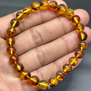 Peut inclure: Un bracelet composé de perles d'ambre. Les perles sont rondes et présentent des nuances de brun et de jaune. Le bracelet est monté sur un cordon élastique jaune. Les perles d'ambre sont translucides.