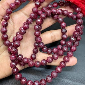 Puede incluir: Un collar de cuentas de piedras preciosas rojo oscuro facetadas. Las cuentas están ensartadas, formando un collar o rosario. Una borla roja cuelga del extremo. Las cuentas tienen un aspecto brillante y pulido.