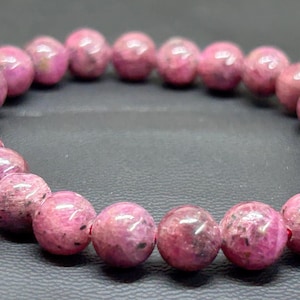 Puede incluir: Una pulsera de cuentas circular compuesta por numerosas piedras esféricas pulidas en varios tonos de rosa intenso, magenta y rojo oscuro. Cada cuenta presenta una superficie lisa y brillante con inclusiones naturales y motas oscuras.