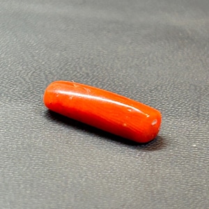 May include: A bright red, cylindrical gemstone, possibly coral, with a smooth, polished surface. The gemstone is approximately 3.8 cm long and lies on a dark, textured surface. The color is a bright, solid red.