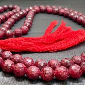 May include: A necklace of faceted, deep red beads with a matching red tassel. The beads are arranged in multiple strands, creating a layered effect. The necklace is displayed on a dark surface.