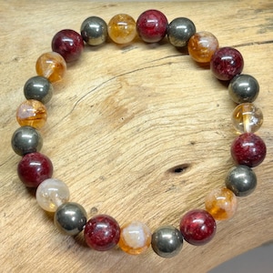 Könnte beinhalten: Ein Armband mit bunten Edelsteinperlen. Es besteht aus runden Perlen in Rot-, Gold- und Grautönen mit klaren und undurchsichtigen Texturen. Das Armband liegt auf einer Holzoberfläche.