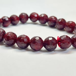 Puede incluir: Una pulsera de cuentas con piedras preciosas facetadas de color rojo intenso. Las cuentas redondas están ensartadas, creando un bucle continuo. La pulsera se muestra sobre un fondo blanco, resaltando el color y la textura de las piedras.