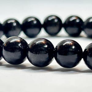 Puede incluir: Primer plano de una pulsera de cuentas negras. Las cuentas son redondas, brillantes y de tamaño uniforme. La pulsera está ensartada en un cordón negro, y las cuentas reflejan la luz.