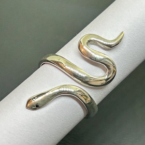 Peut inclure: Une bague serpent argentée. La bague s'enroule autour du doigt, la tête et la queue du serpent se rejoignant. La tête du serpent a un petit œil en pierre noire. La bague est présentée sur une surface blanche.