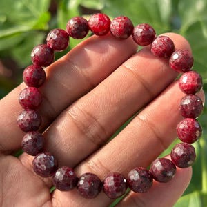 May include: A bracelet made of faceted, deep red gemstones. The round, polished beads are a rich, dark red colour. The beads are strung together, forming a complete circle. The bracelet is shown against a blurred green background.