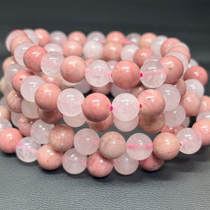 Könnte beinhalten: Nahaufnahme mehrerer Armbänder aus rosa und weißen Edelsteinperlen. Die runden Perlen sind auf einem rosa elastischen Band aufgereiht. Die Armbänder sind gestapelt und zeigen die verschiedenen Rosa- und Weißtöne.