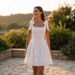 以下が含まれることがあります： スクエアネックとリボンストラップが付いた白いドレス。 ドレスはフィットしたボディスとフレアスカートです。 女性は笑顔で、石の壁と緑の植物の前で屋外に立っています。