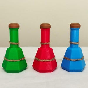 Könnte beinhalten: Drei farbige Dekoflaschen. Die Flaschen sind grün, rot und blau, jede mit einem braunen Deckel und einem Bindfadenband. Die Flaschen haben ein geometrisches, facettiertes Design.