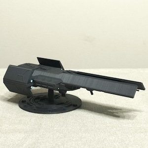 Op de afbeelding: Een zwart model van een futuristisch ogende geschutstoren. Het model is gemonteerd op een cirkelvormige basis met een gedetailleerd ontwerp. Het kanon heeft een lange, rechthoekige loop en een boxy hoofdgedeelte. Kleine blauwe lampjes zijn zichtbaar.