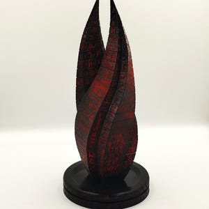 May include: A tall, stylized sculpture with a black base and a red and black upper section. The upper section has a textured surface with intricate patterns and rises to two sharp points. The sculpture is set against a white background.