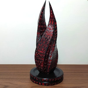 Puede incluir: Una escultura negra en forma de doble hélice con intrincados grabados rojos en relieve por toda su superficie. El objeto se alza verticalmente sobre una base circular negra, también decorada con símbolos grabados en color rojo.