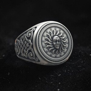 May include: A silver signet ring featuring a circular face with a raised relief of a sun face surrounded by rays. The band is decorated with intricate, symmetrical knotwork patterns on the sides, finished with a dark, oxidized silver patina.