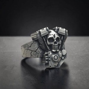 May include: A silver-toned ring featuring a detailed skull integrated into a V-twin engine block design. The band displays a cracked, weathered texture, while the engine component includes a small circular emblem with a skull icon at the center.