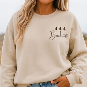 Puede incluir: Sudadera beige con los números "444" encima de la palabra "Beschützt" en escritura cursiva. La sudadera es de color neutro y tiene un corte holgado.
