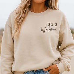 Puede incluir: Sudadera beige de cuello redondo con el número "555" y la palabra "Wachstum" impresa en negro. La sudadera se combina con vaqueros azules. El estilo es informal y cómodo.