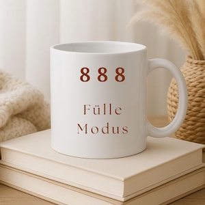 Può includere: Tazza in ceramica bianca con il numero "888" e le parole "Fülle Modus" in marrone. La tazza è appoggiata su due libri impilati. Sullo sfondo si vedono un cestino intrecciato e piante secche.
