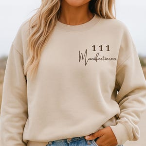 Puede incluir: Sudadera beige de cuello redondo con el texto "111 Manifestieren" en escritura negra. La sudadera es de color neutro y tiene un corte relajado. La modelo lleva vaqueros azules.