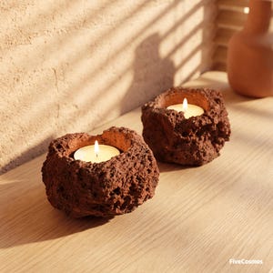 May include: Two tea light candle holders made from porous, reddish-brown volcanic rock. Each holder features a rough, uneven exterior texture and a central circular cavity containing a small, lit white tea light candle with a warm yellow flame.