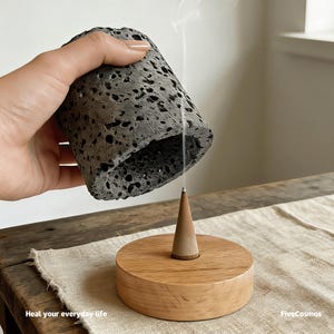 Może przedstawiać: Szary, porowaty uchwyt na kadzidło jest trzymany nad drewnianym stożkiem i podstawą. Dym unosi się ze stożka, wskazując na palące się kadzidło. Podstawa jest z jasnego drewna, a uchwyt ma teksturowany, wulkaniczny wygląd. Tekst "Heal your everyday life" znajduje się na dole.