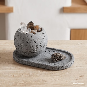 May include: A grey, porous, spherical concrete bowl resting on an oval tray of the same material. The bowl is filled with various clear and brown crystals. A small, porous, grey stone fragment sits on the tray. The text FiveCosmos is visible.