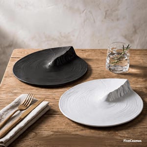 Könnte beinhalten: Ein schwarzer Teller und ein weißer Teller, beide mit einer erhabenen, strukturierten, bergähnlichen Erhebung und konzentrischen kreisförmigen Rillen. Ein Glas mit Wasser und einem Rosmarinzweig sowie eine goldene Gabel und ein Messer liegen bereit.
