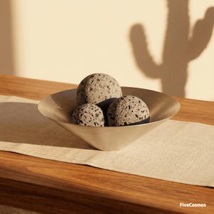 May include: A shallow, brushed metallic bowl with a conical base containing three spherical, porous gray volcanic rocks. The text "FiveCosmos" is printed in the bottom right corner of the image, which is illuminated by warm, directional light.