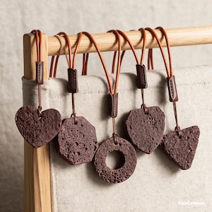 May include: Five dark brown, porous, volcanic rock pendants in heart, octagon, circle, shield, and triangle shapes. Each pendant hangs from a thin, tan leather cord secured with a dark brown leather sleeve. The text FiveCosmos is visible.