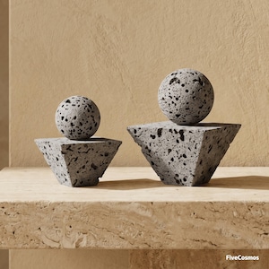 Puede incluir: Dos esculturas decorativas hechas de roca volcánica gris porosa. Cada pieza consiste en una esfera que descansa sobre una base piramidal. Los objetos presentan una textura rugosa y llena de poros. El texto FiveCosmos es visible abajo a la derecha.