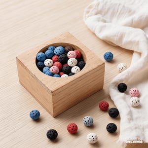 May include: A wooden square container filled with colorful, porous lava rock spheres. The spheres are in various colors, including blue, red, black, white, and pink. Some spheres are scattered around the container on a light wooden surface. A white cloth is in the background.