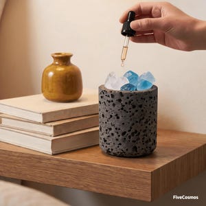 Pode incluir: Um difusor de óleos essenciais de pedra porosa cinza escuro com cristais azuis e brancos. Um pequeno vaso amarelo mostarda e uma pilha de livros estão por perto. Um conta-gotas dispensa óleo no difusor. O texto "FiveCosmos" está no canto inferior direito.