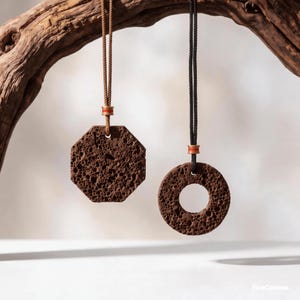 Peut inclure: Deux pendentifs en roche volcanique poreuse brun foncé. L'un est octogonal et suspendu à un cordon beige, l'autre est circulaire avec un trou central et suspendu à un cordon noir. Chaque cordon comporte une perle dorée avec une rayure rouge.