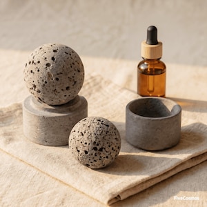 May include: Two porous gray stone spheres, one resting on a cylindrical concrete base and one sitting beside a small concrete bowl. An amber glass dropper bottle with a wooden cap stands nearby. The text FiveCosmos is visible in the corner.